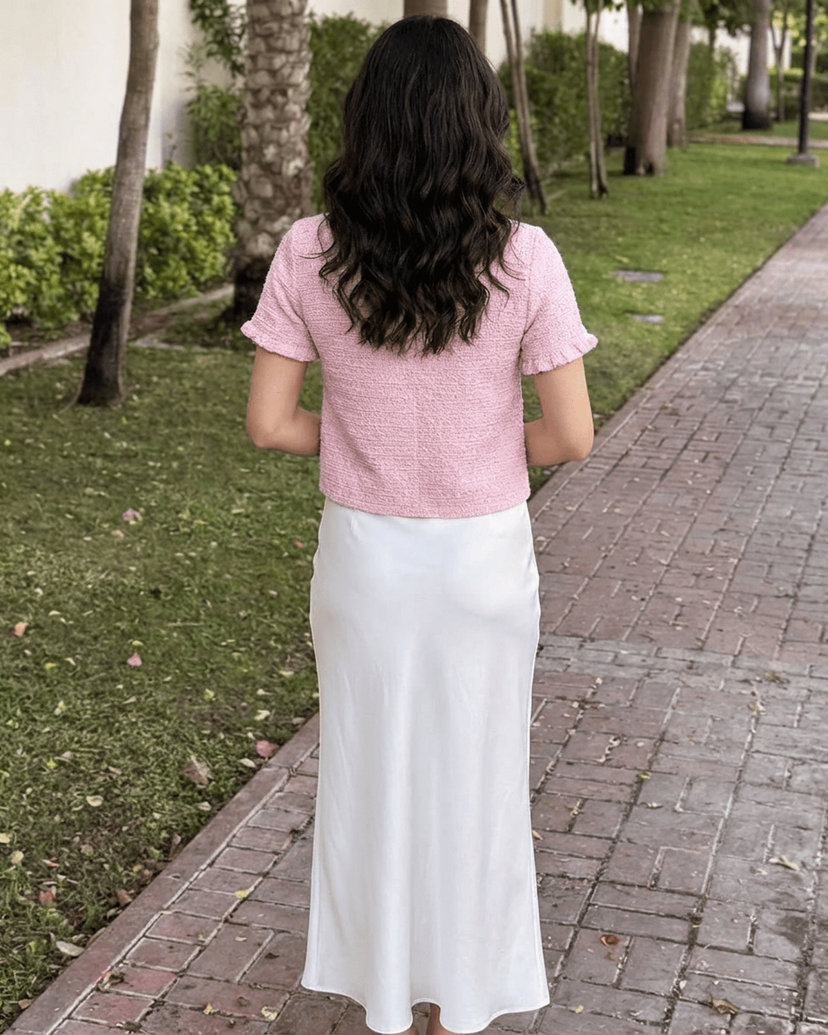 Back view of woman in Camellia Cardigan by Lauven, styled with a white skirt in a garden setting.