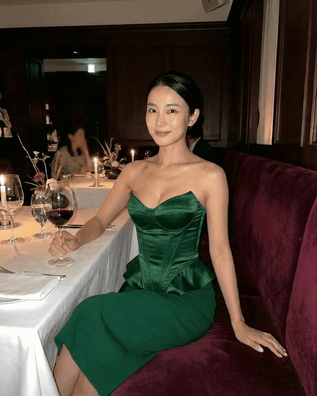 Model wearing Lauven Emerald Corset Top, a strapless satin top in deep green, seated at a dining table.