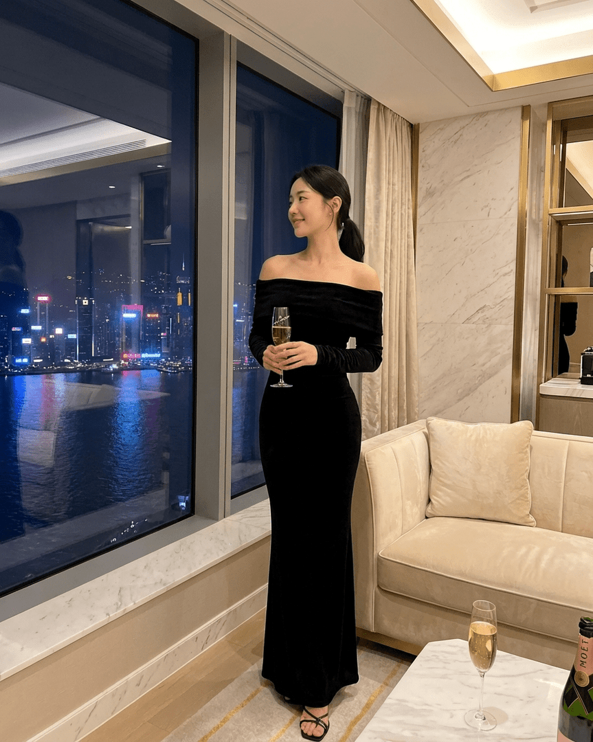Elegant woman in Noctura Maxi Dress by Lauven, off-shoulder design, standing by a window with city view.