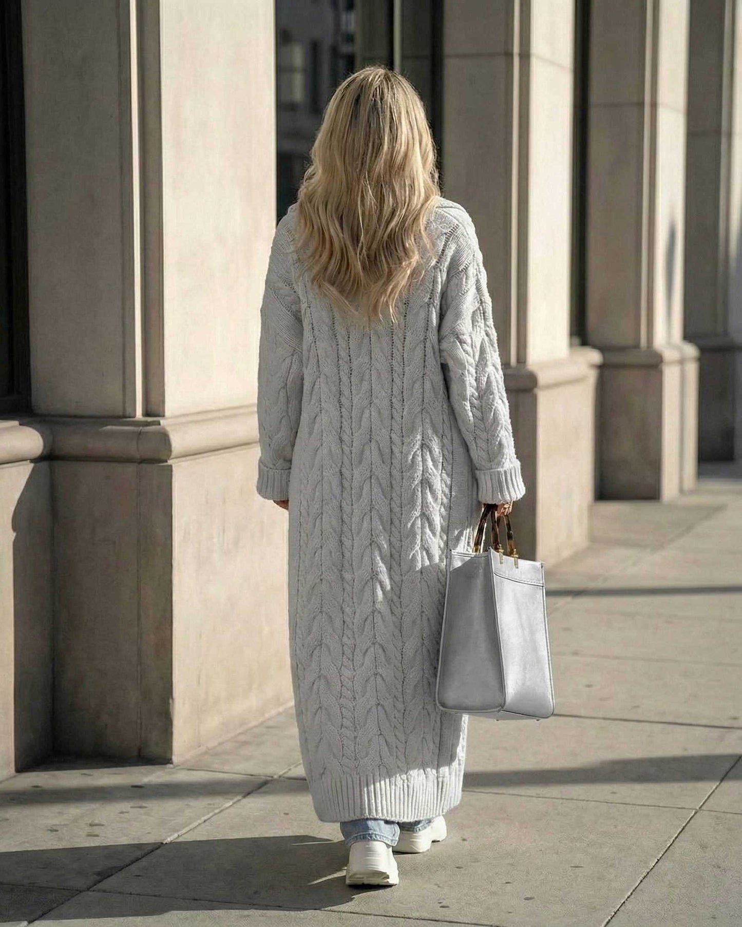 Woman wearing a long cable-knit Verona Cardigan in grey, walking with a tote bag in a city setting.