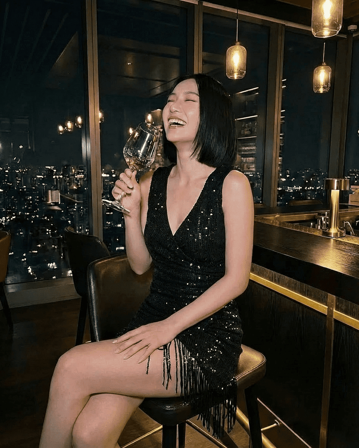 Lauven Noir Mini Dress worn by a woman laughing while holding a wine glass, featuring a black sequin design and fringe hem.