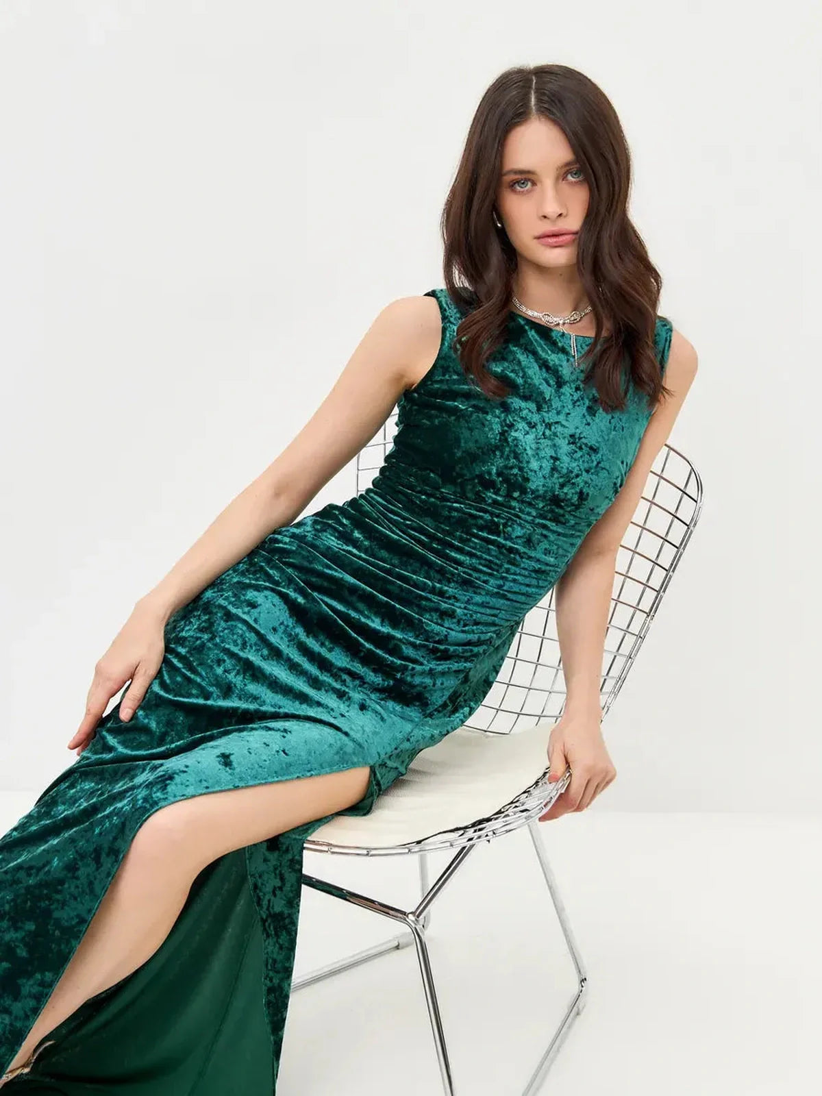 Woman in a sleek green Velvet Aura Maxi Dress with ruching and a high slit, seated on a modern chair.