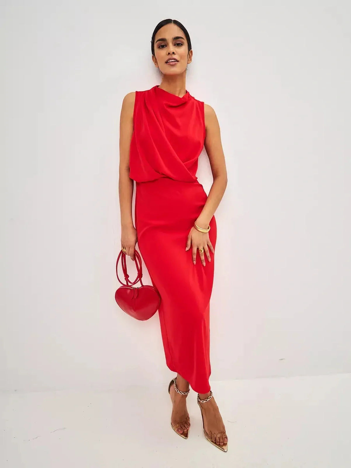 Stylish woman wearing a sleeveless red drape maxi dress with heart-shaped handbag, showcasing a polished evening look.