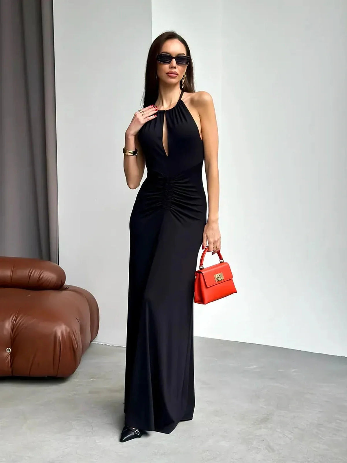 Elegant black maxi dress with halter neckline, keyhole cutout, and front ruching, styled with red handbag and sunglasses.