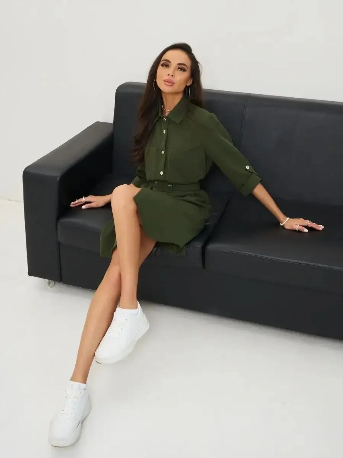 Model showcasing an olive green belted shirt mini dress, styled with white sneakers, seated on a black couch.