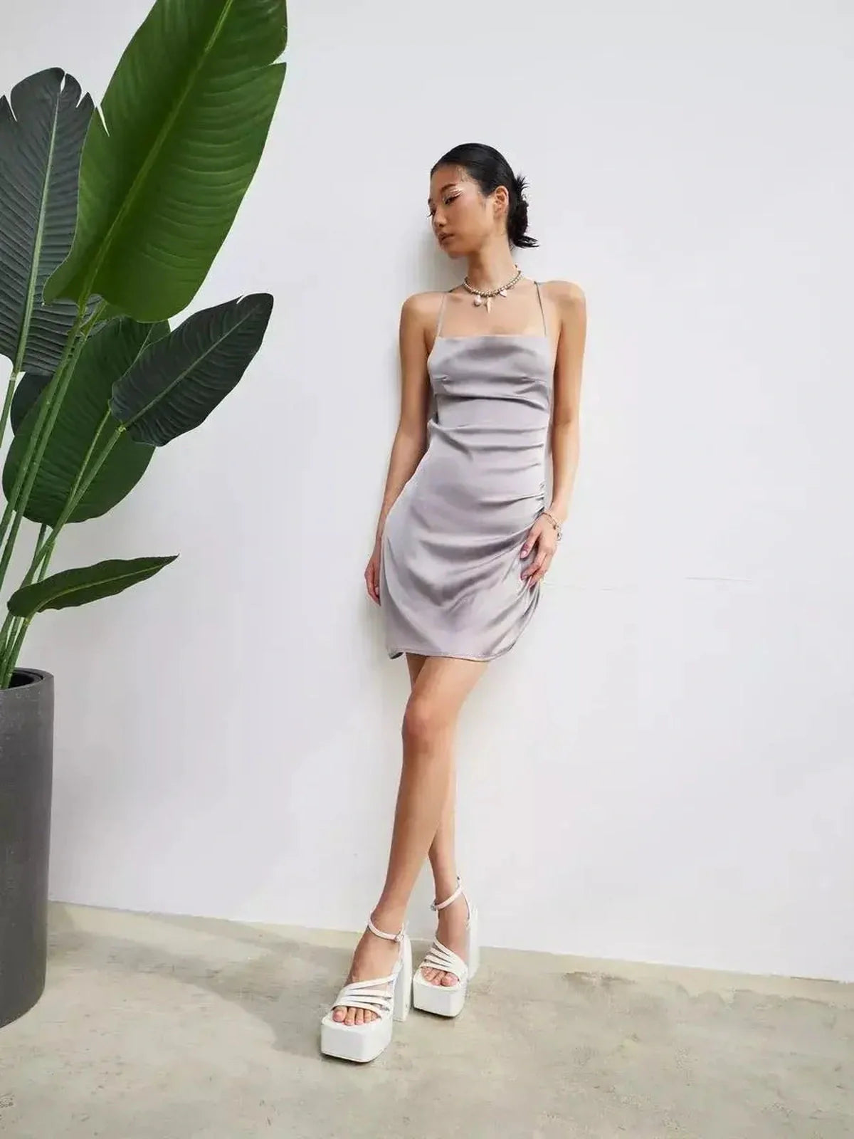 Model wearing a silver-gray satin mini dress with crisscross tie straps, posing against a minimalist background.