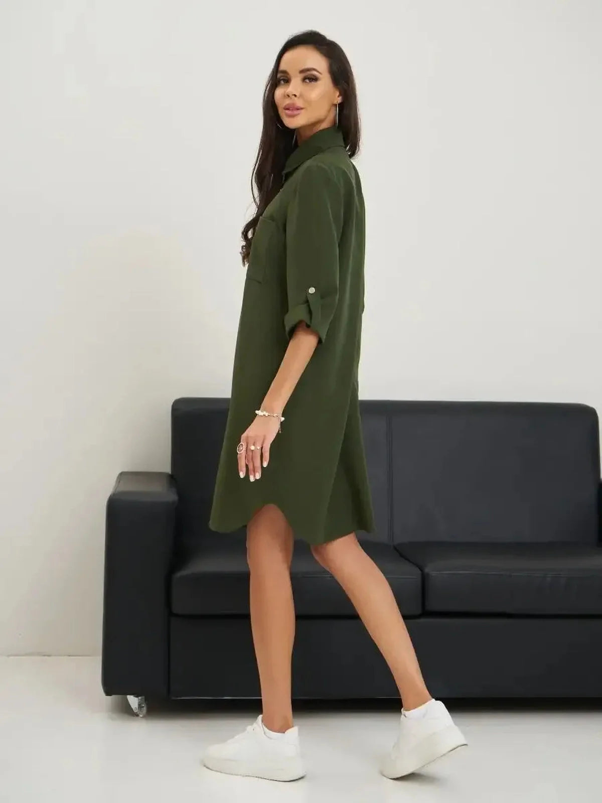 Model wearing an olive green belted shirt mini dress styled with sneakers against a modern couch backdrop.