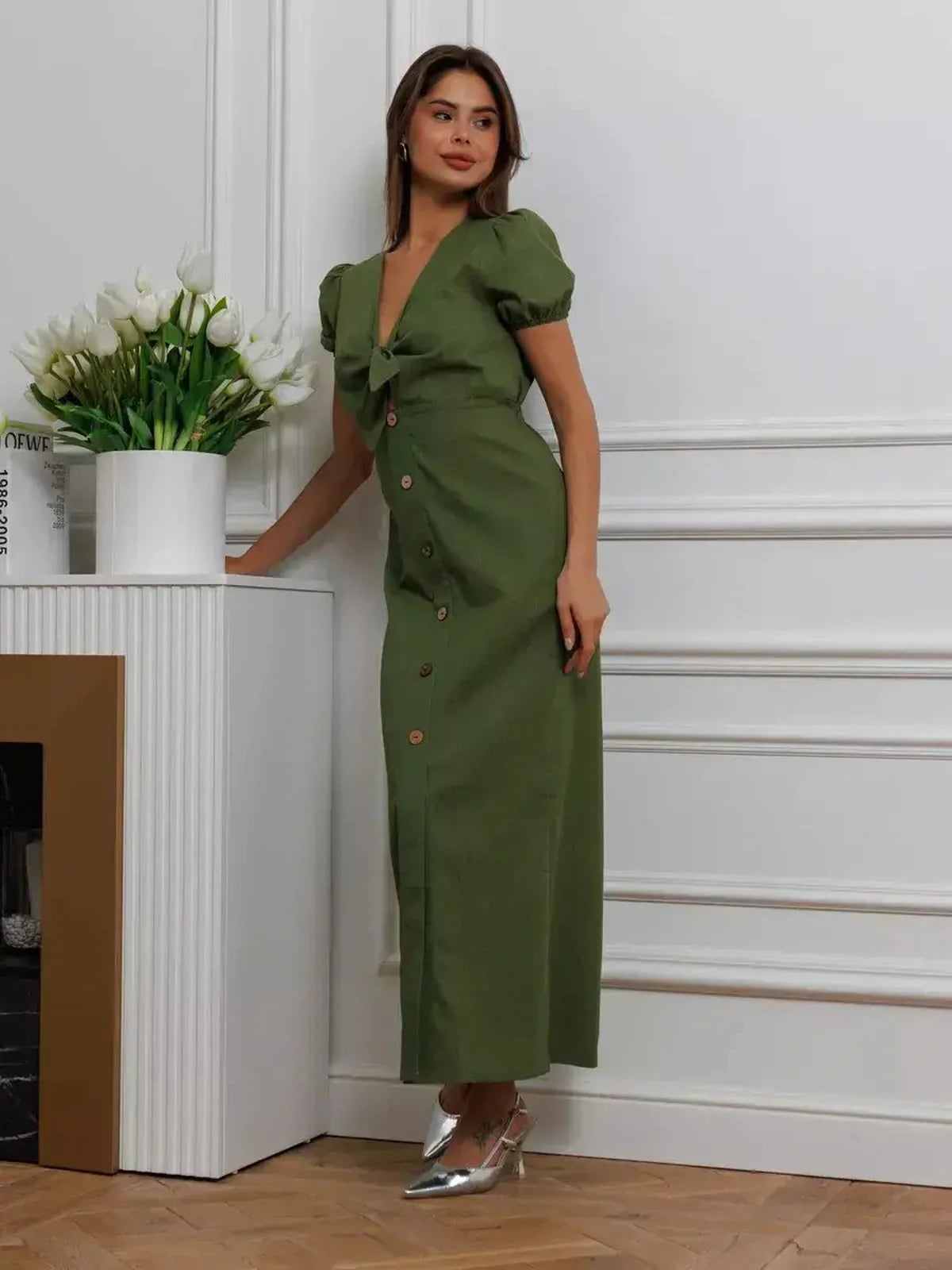 Model wearing Olive Whisper Maxi Dress in khaki with puff sleeves and tie front, standing beside a white vase with flowers.