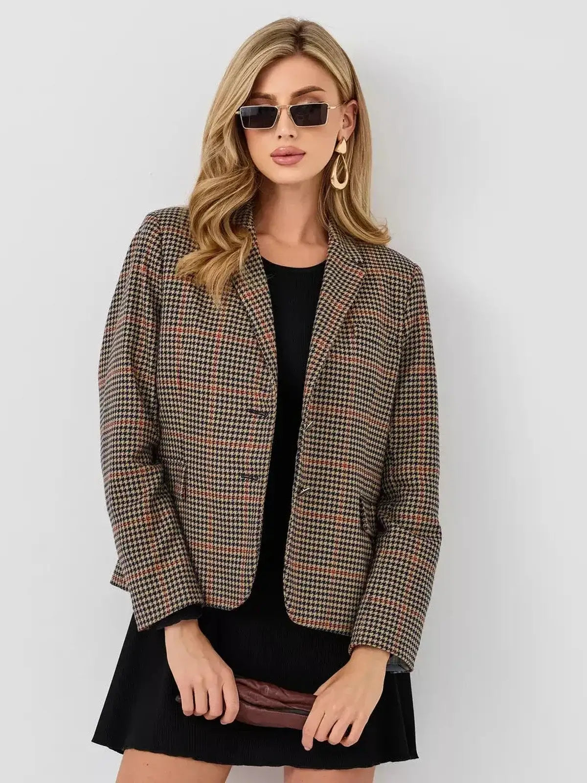 Chic woman in plaid blazer over black dress, accessorized with stylish sunglasses and clutch for a modern look.