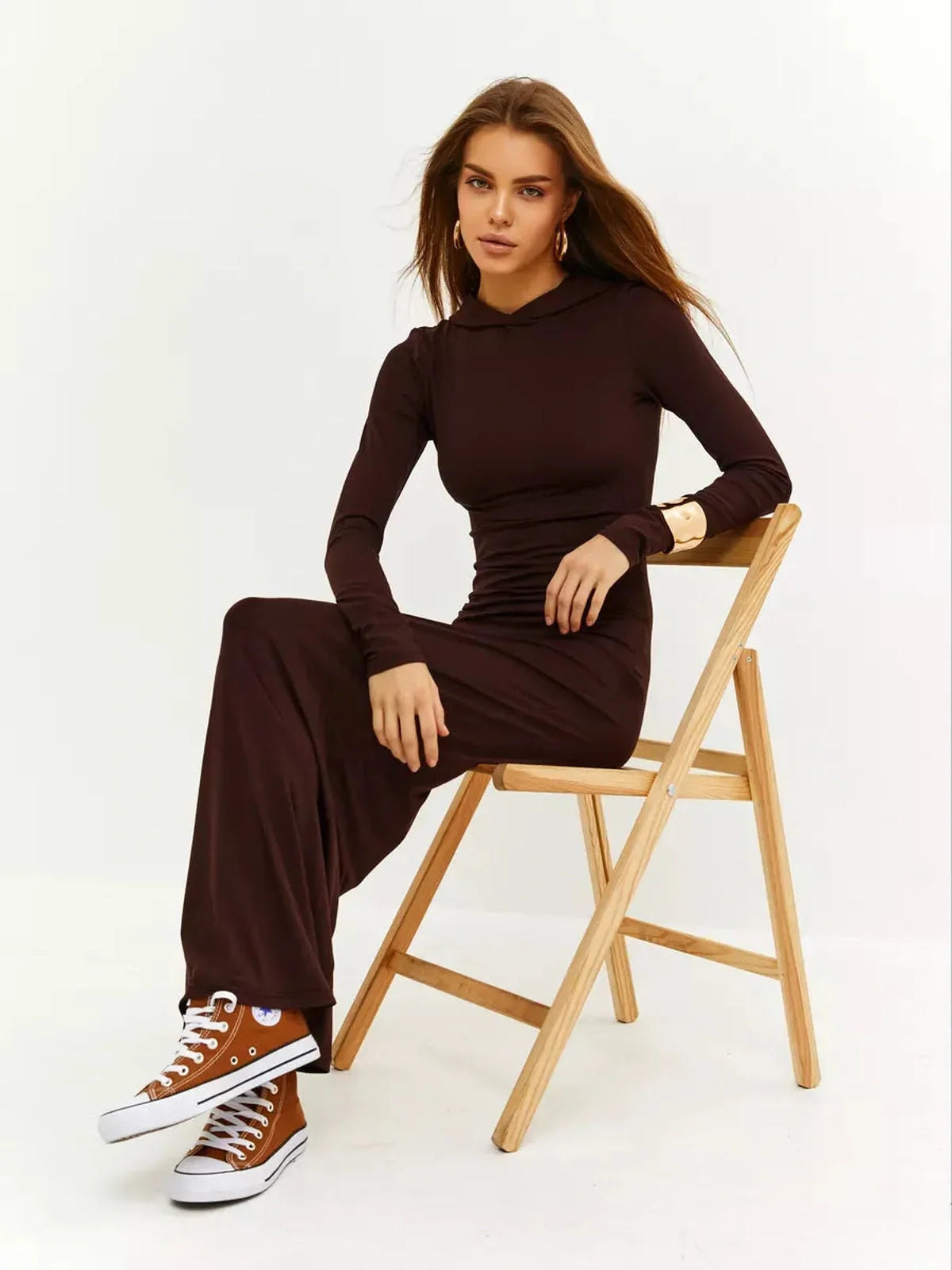 Model in a chocolate-toned Sculpt Maxi Dress with hood, seated on a chair, styled with sneakers.