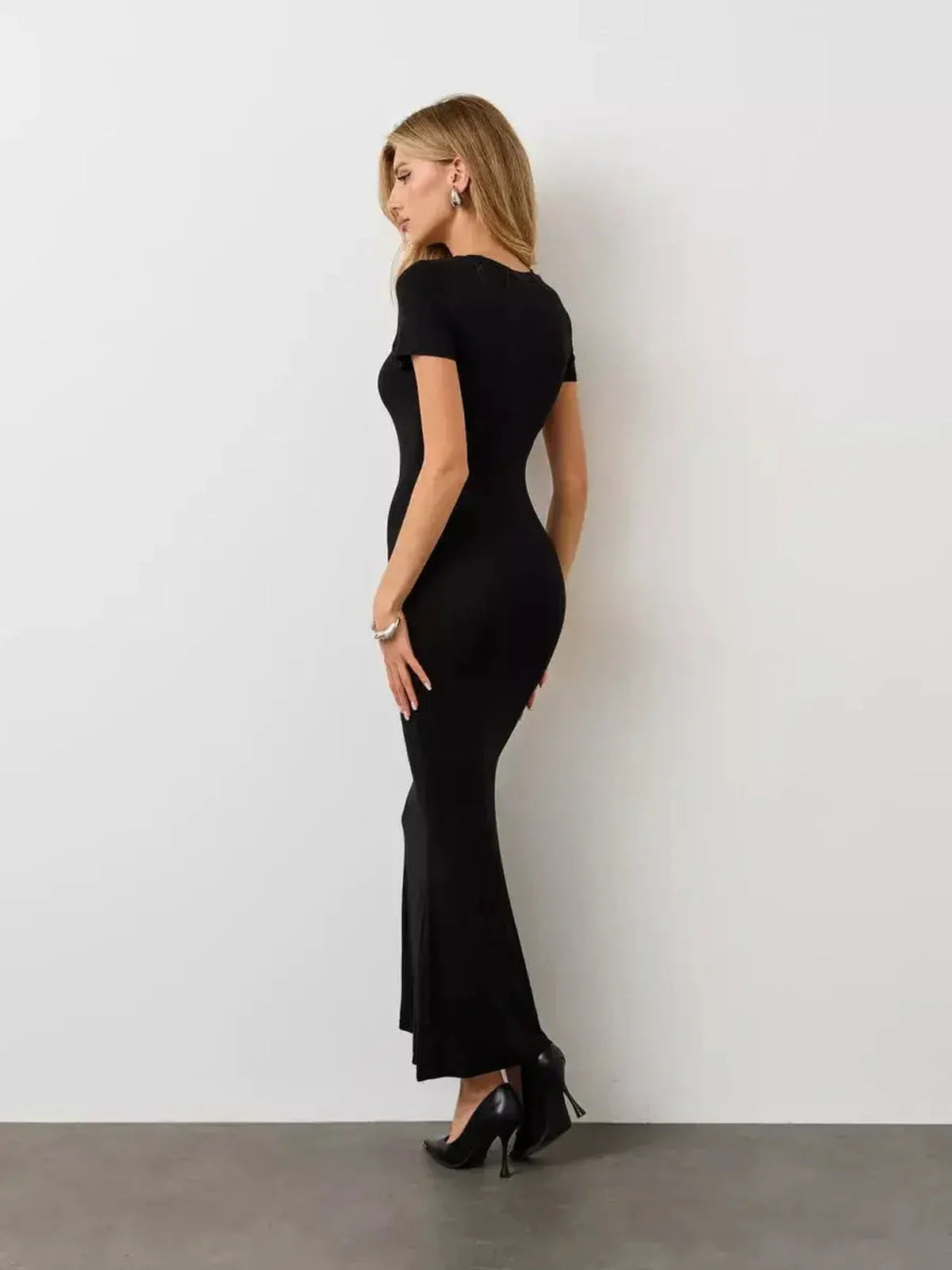 Elegantly styled woman in a Noir Curve Maxi Dress showcasing its sculpted silhouette and flared hem against a minimalist backdrop.