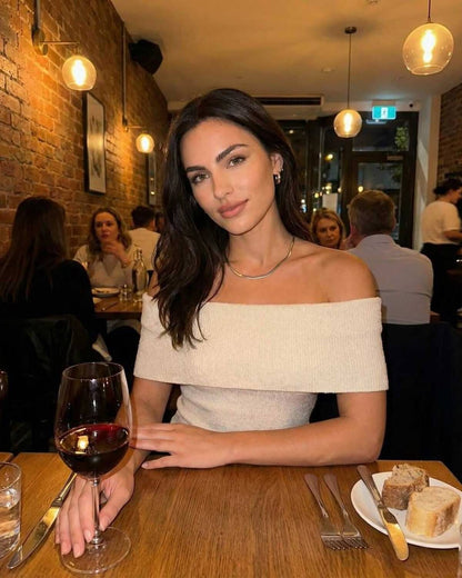 Model wearing Lauven Monaco Top in a cozy restaurant setting, showcasing elegant off shoulder design with wine.