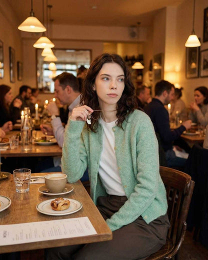 Sage Cardigan by Lauven worn by a woman in a cozy café setting, showcasing its stylish round neck and button front.