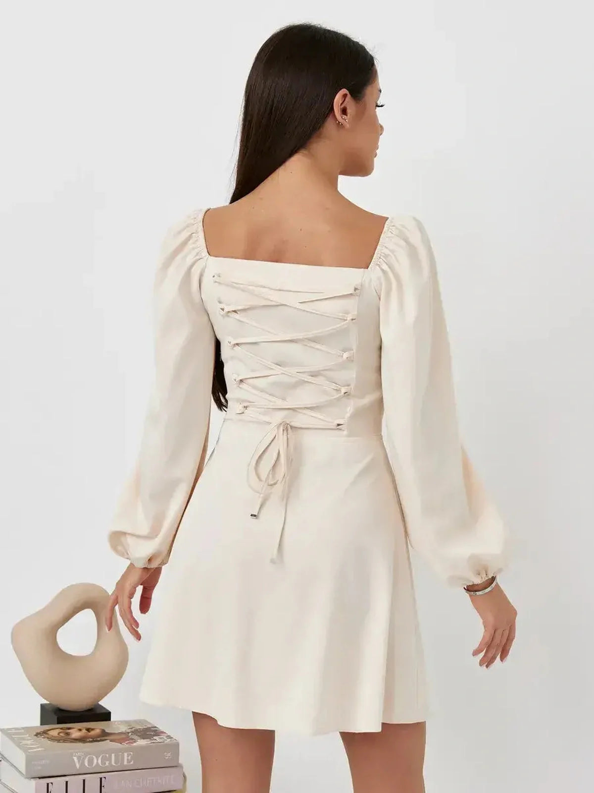 Back view of a woman in a beige Lace Muse mini dress with a square neckline and lace-up bodice details.