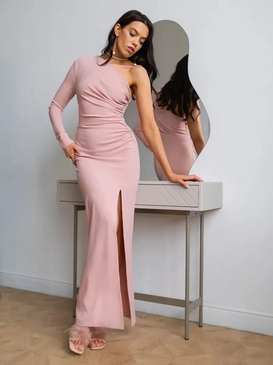 Model wearing a one-shoulder maxi dress in blush with draped details and a front slit, styled elegantly against a mirror.
