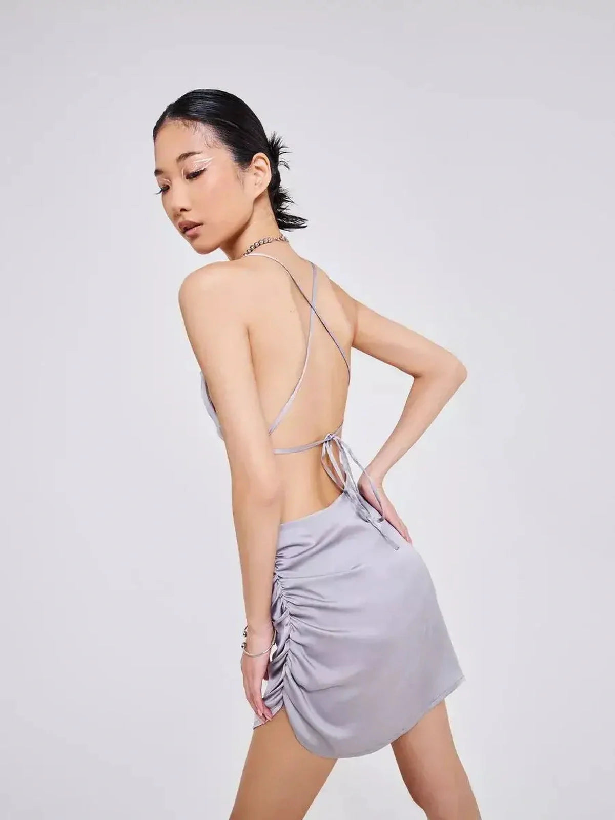 Back view of a model wearing the Silken Rush silver-gray satin mini dress with crisscross tie straps and side ruching.