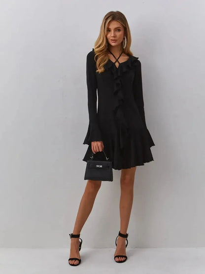 Model wearing Velvet Noir black midi dress with ruffles and V-neckline, holding a small handbag and displaying stylish heels.