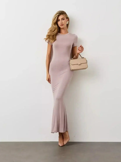 Bare Essence Maxi Dress in beige worn by a model with a stylish handbag, showcasing the flared hem and curve-tracing fit.
