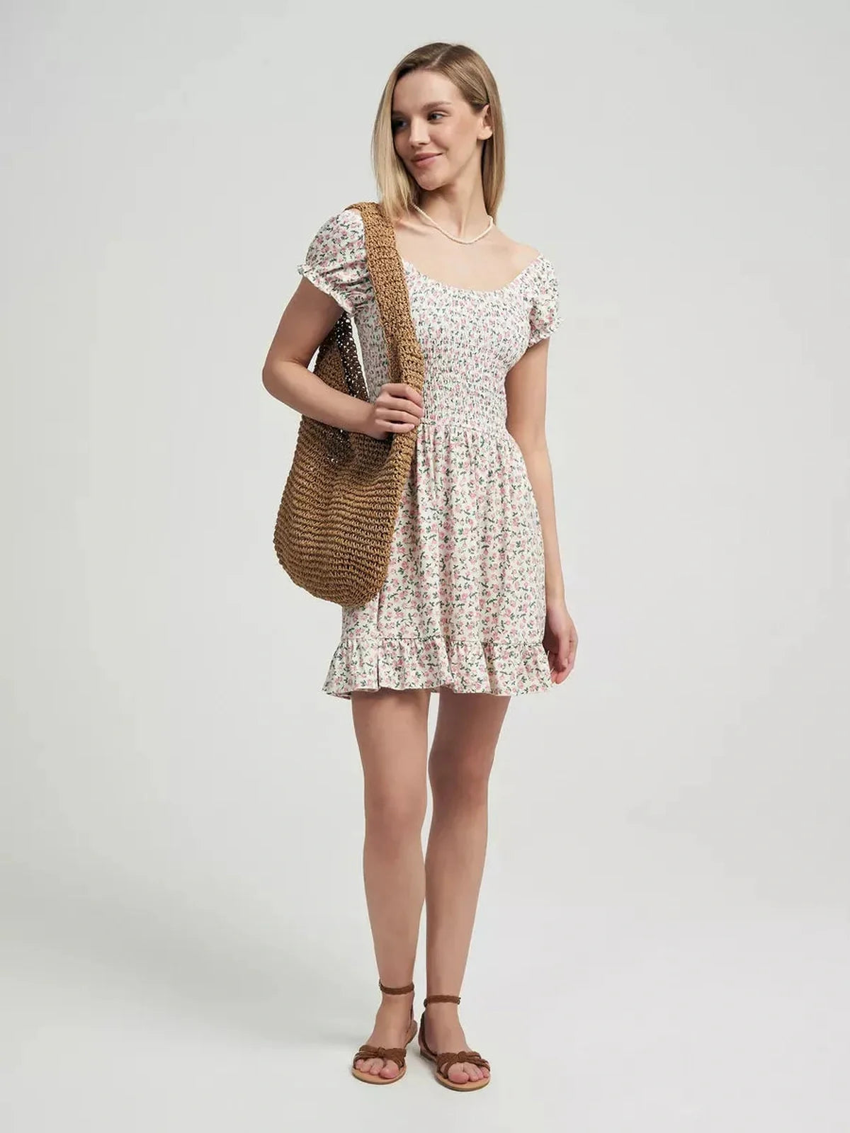 Model wearing the Blush Blossom summer dress with floral prints and a ruffled hem, styled with a woven bag.