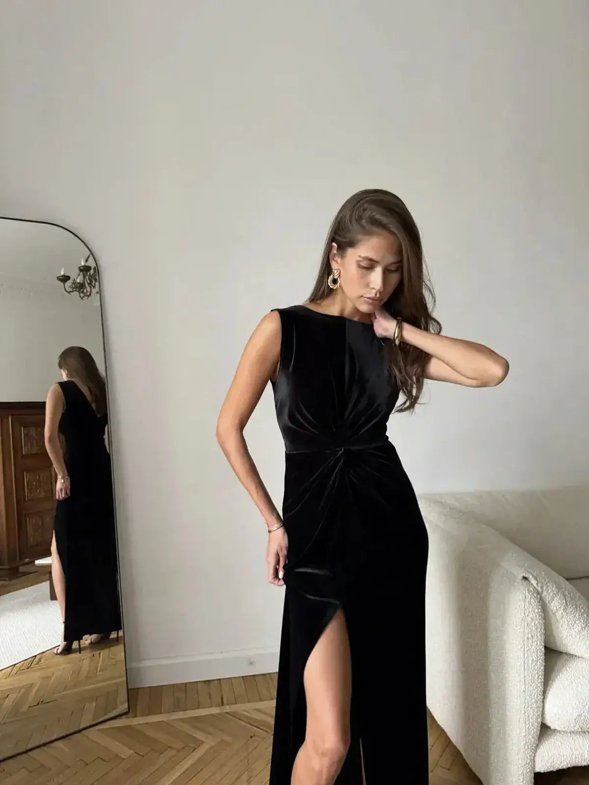 Model showcasing the Eclipse Maxi Dress in black velvet, featuring a draped knot and thigh-high slit against a stylish interior.