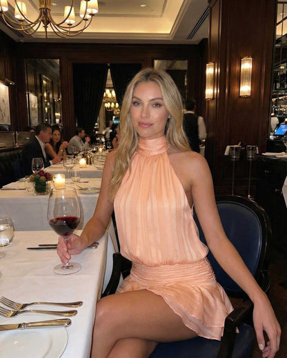 Model wearing Aprielle Mini Dress by Lauven, a pleated chiffon halter mini dress in peach, seated at a stylish restaurant.