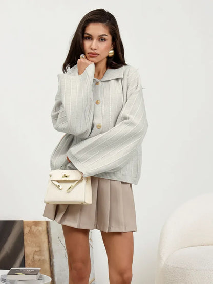 Chic woman wearing Glacier Cardigan in light gray with oversized sleeves and braided detail, styled with a pleated skirt.