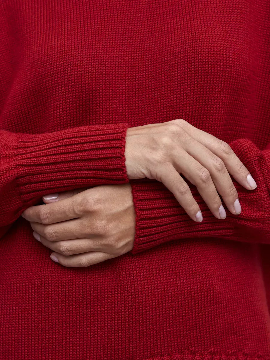 Close-up of hands wearing the Sienna Sweater by Lauven, showcasing its deep red knit turtleneck and ribbed cuffs.