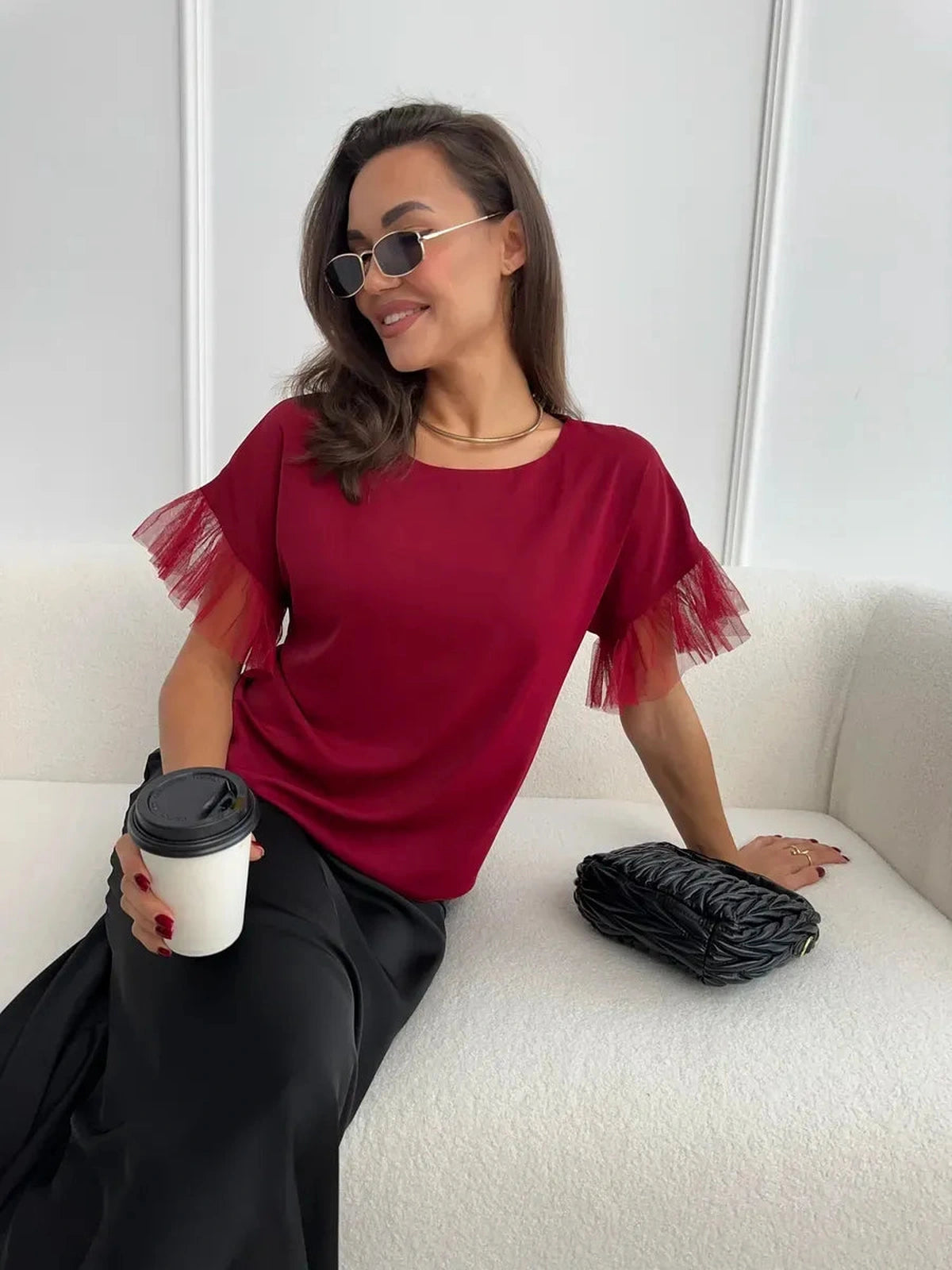 Woman in burgundy Velora Blouse with sheer ruffle sleeves, holding a coffee cup, exuding elegance and style.