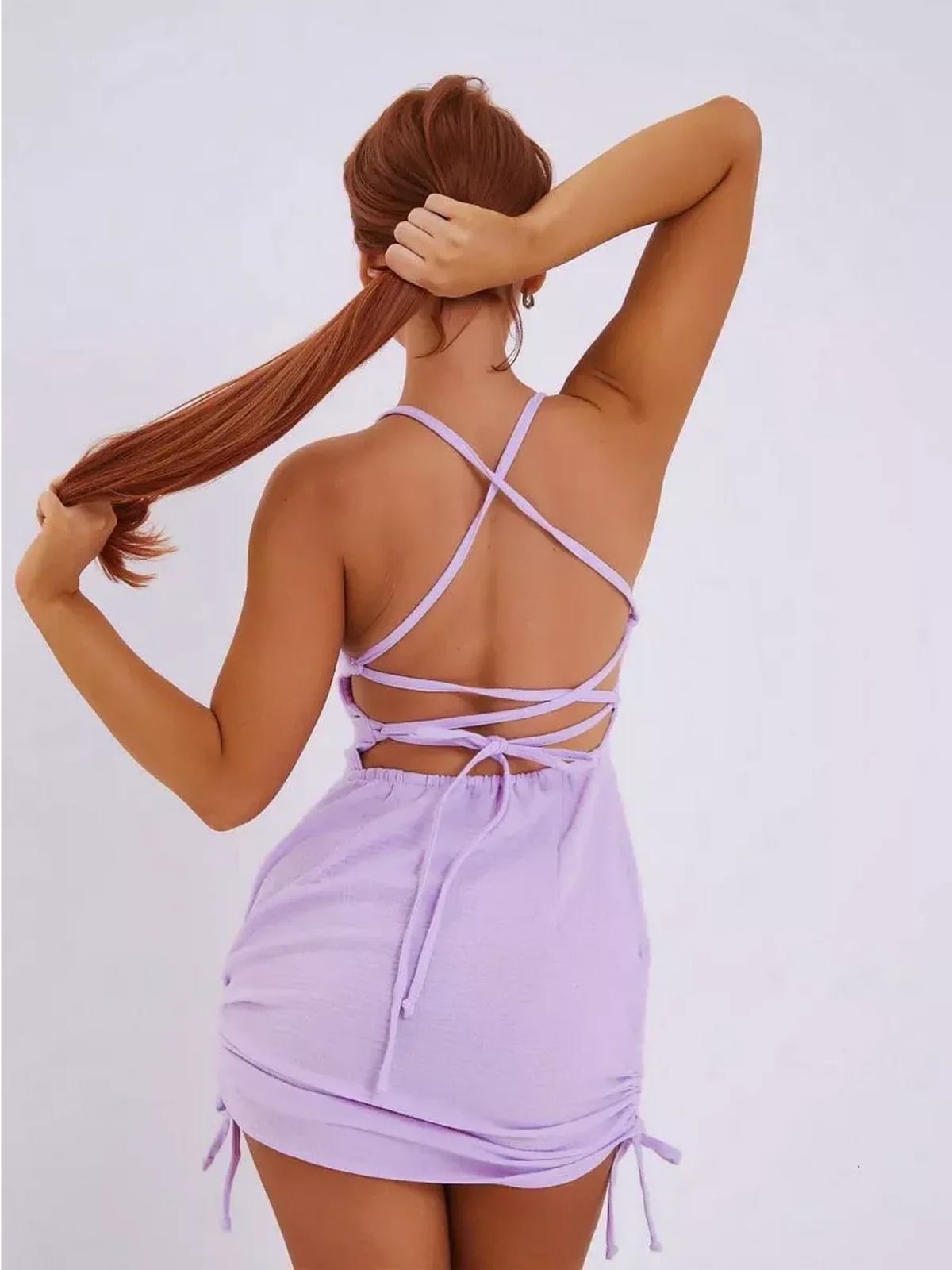 Back view of a woman wearing a lilac mini dress with adjustable criss-cross straps and a ruched hem.