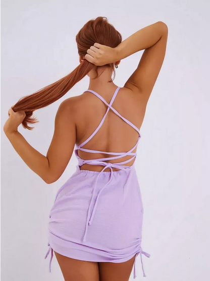 Back view of a woman wearing a lilac mini dress with adjustable criss-cross straps and a ruched hem.