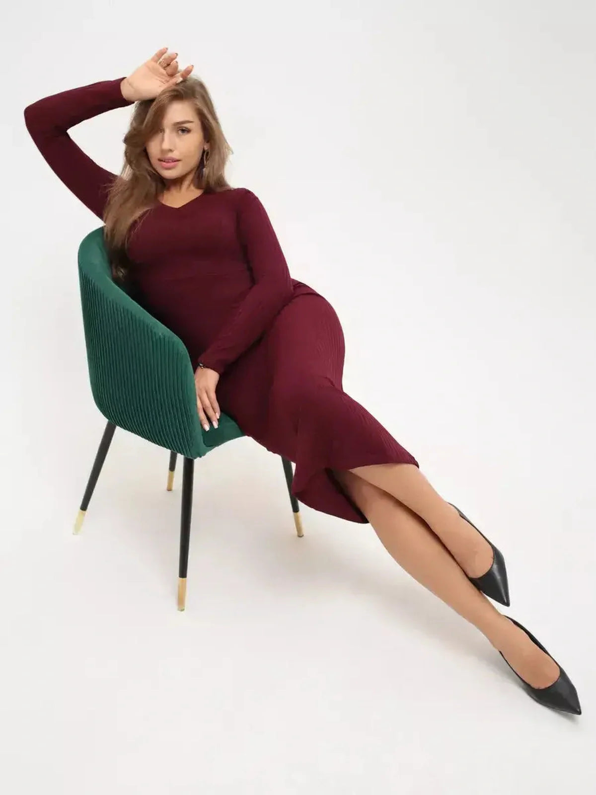 Model showcasing the Merlot Grace Midi Dress, a sleek, body-contouring garment in deep burgundy, seated elegantly on a green chair.