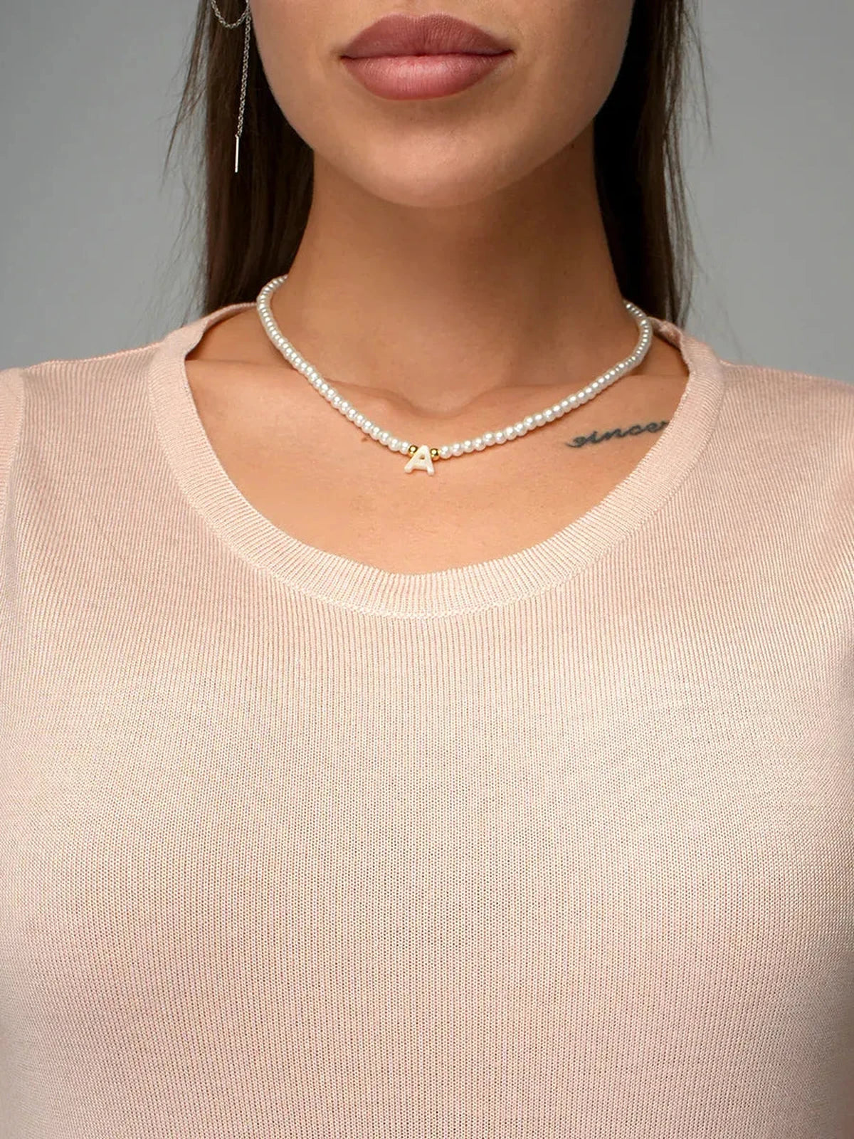 Close-up of a woman wearing a pearl necklace with an 'A' pendant, styled with a beige top.