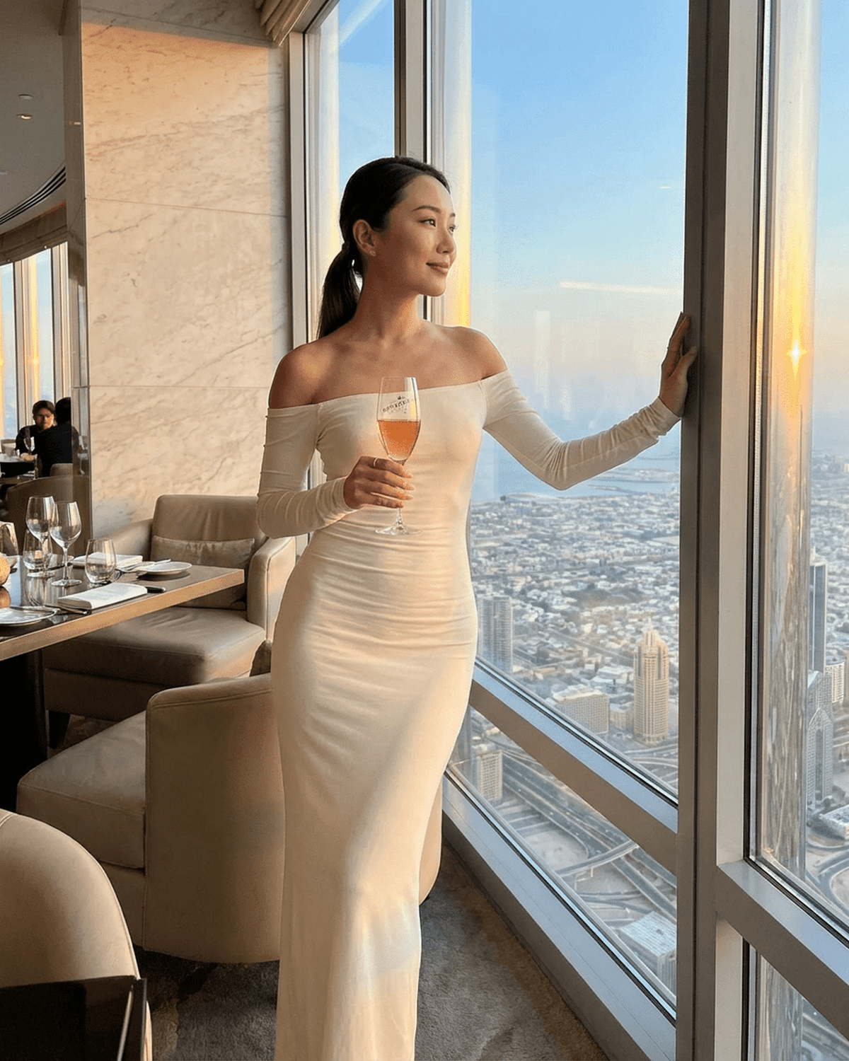 Elegant woman in an Ivory Maxi Dress by Lauven, enjoying a drink while gazing out at the sunset view.