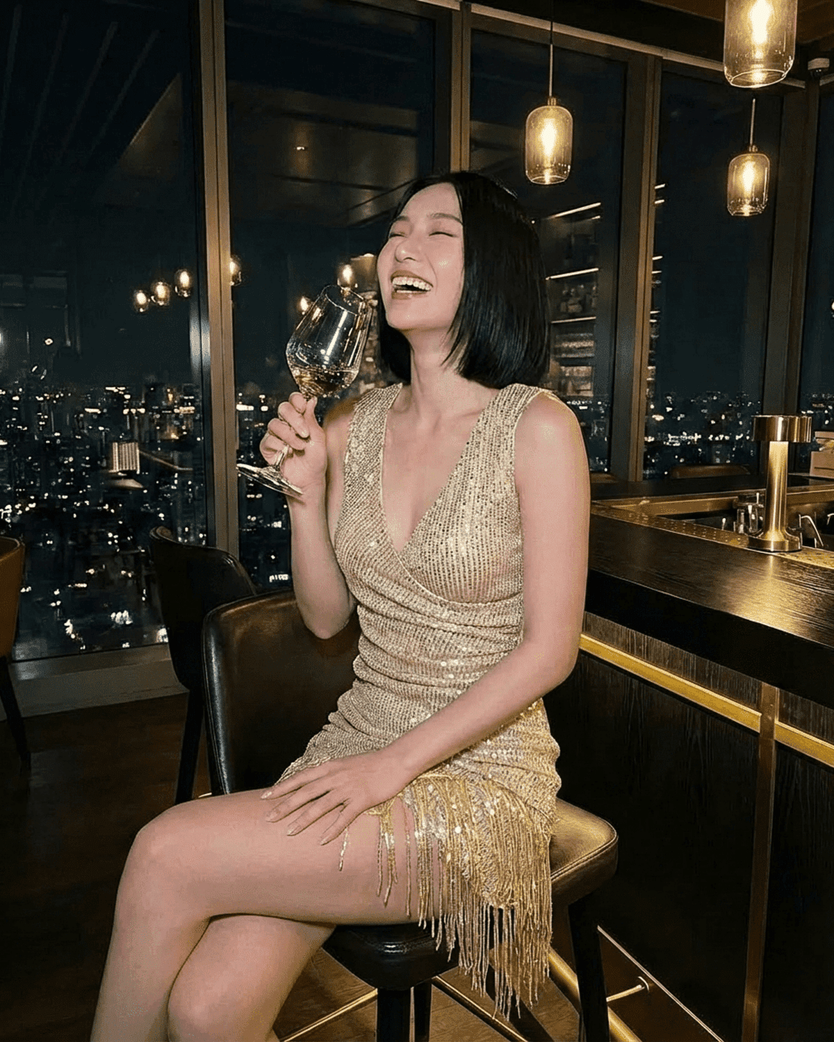A woman laughing in an Aurora Mini Dress by Lauven, featuring gold sequins and fringe hem, holding a glass of wine.
