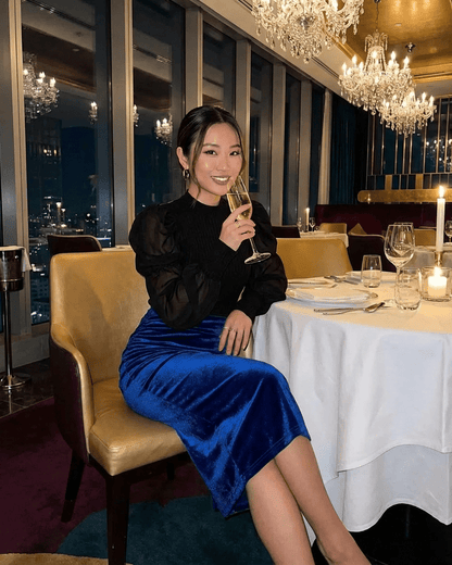 Woman wearing Azure Midi Skirt by Lauven, enjoying a drink at an elegant restaurant setting.