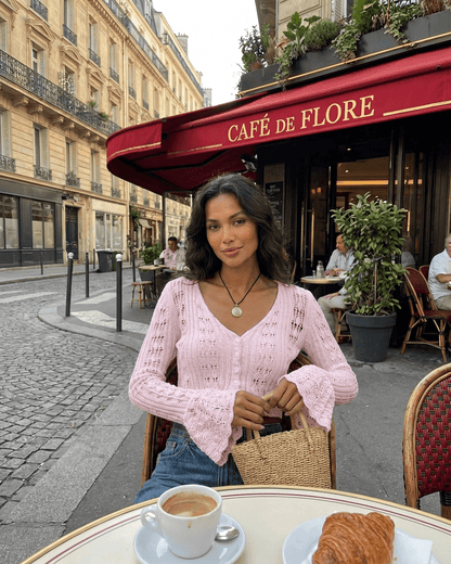 A woman wearing the Blossom Cardigan in pink at Café de Flore, enjoying coffee and a pastry, showcasing style and elegance.