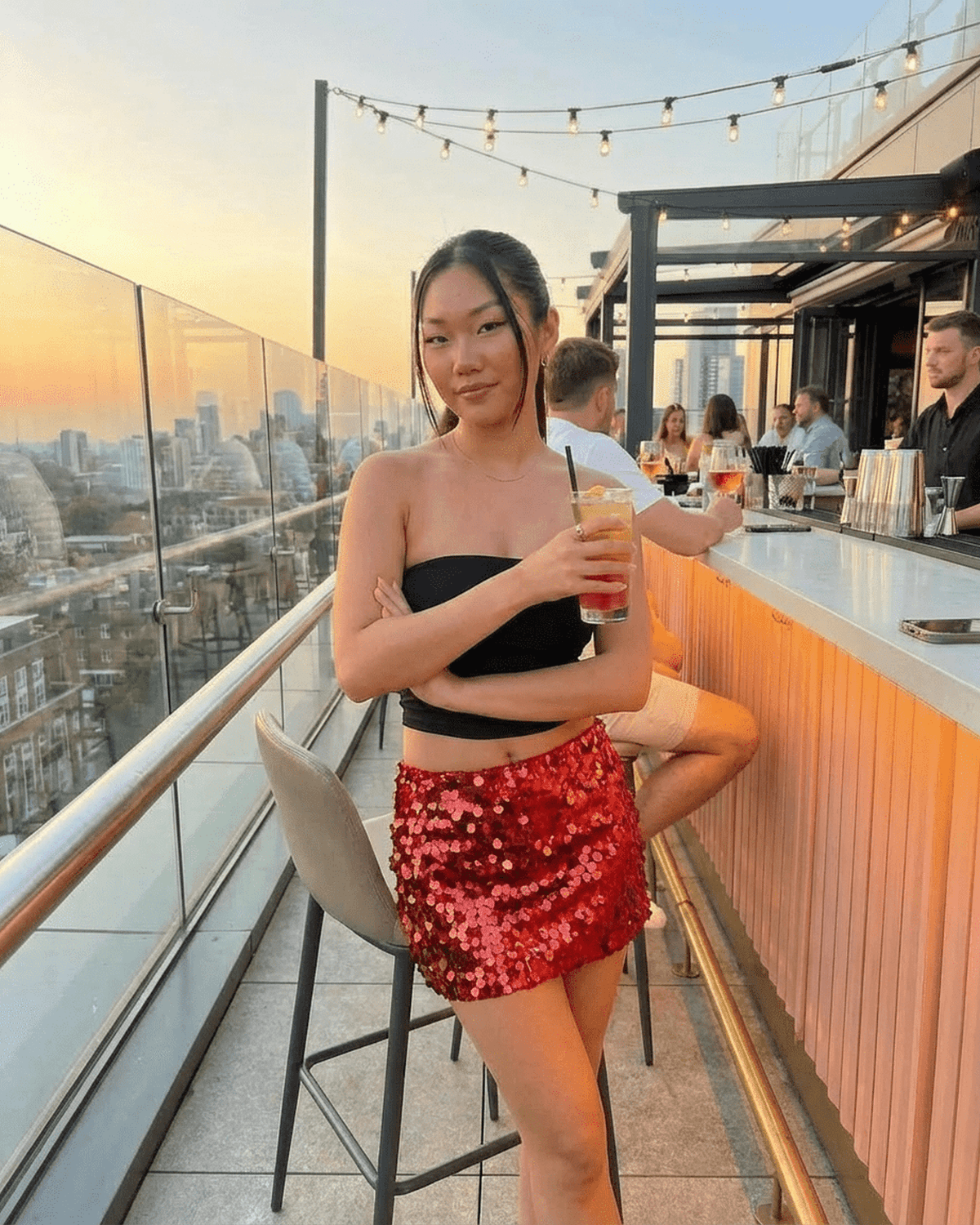 Model wearing Lauven Flamenco Mini Skirt with red sequins, enjoying a cocktail at a rooftop bar during sunset.