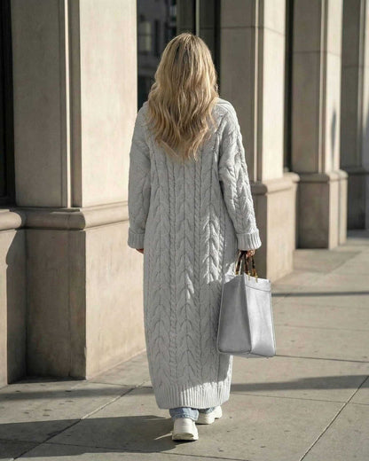 Woman wearing a long cable-knit Verona Cardigan in grey, walking with a tote bag in a city setting.