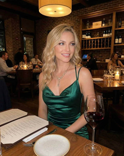Woman wearing Verona Mini Dress by Lauven in emerald green satin, dining elegantly in a restaurant with wine.