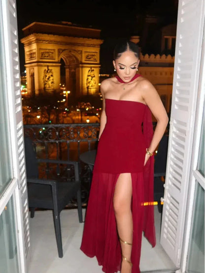 Stunning woman in a red maxi dress with a thigh-high slit, posed elegantly at a balcony with a city view.