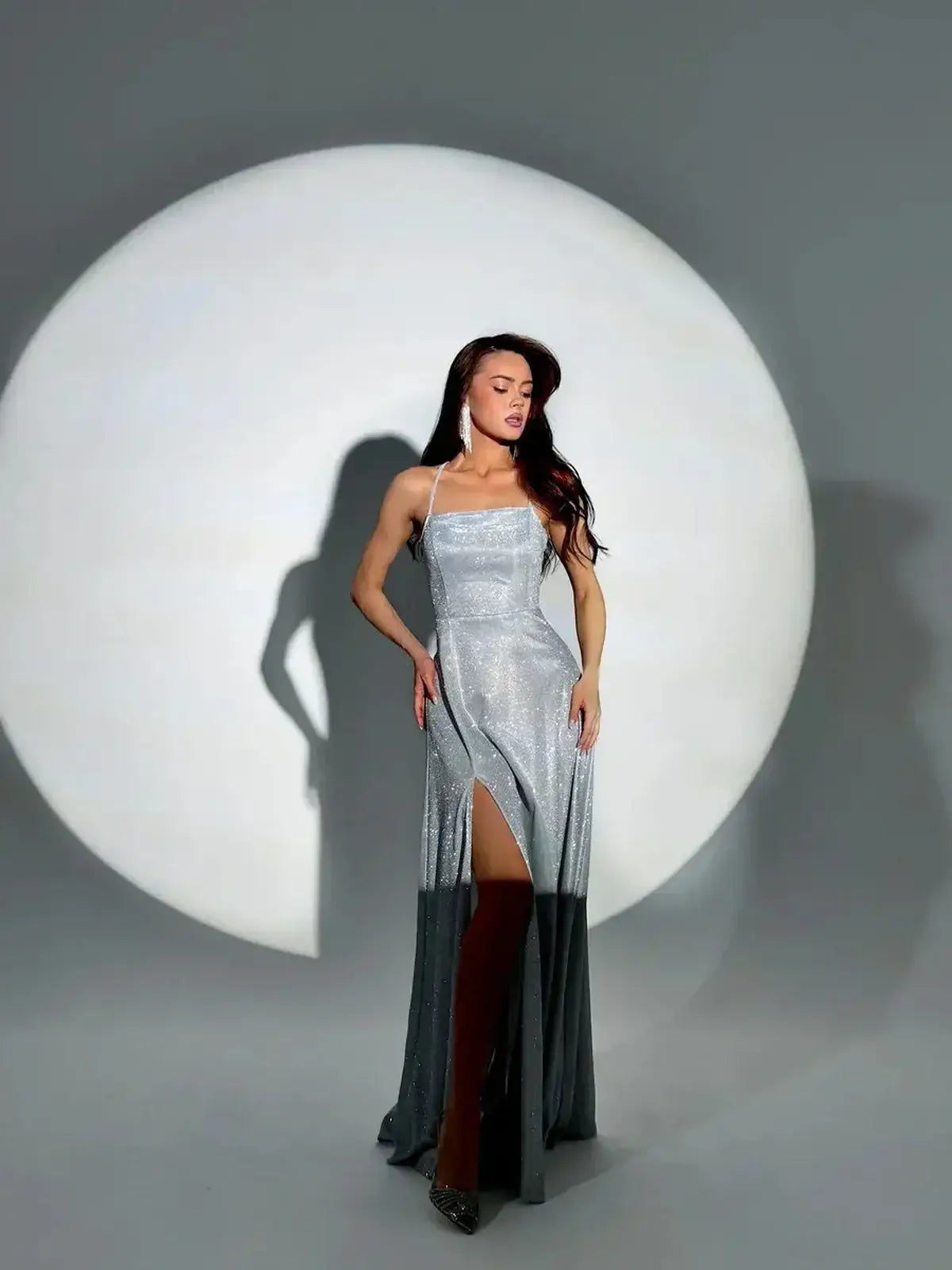 Model in sparkling silver Lunar Halo Maxi Dress with side slit, set against a soft circular backdrop.
