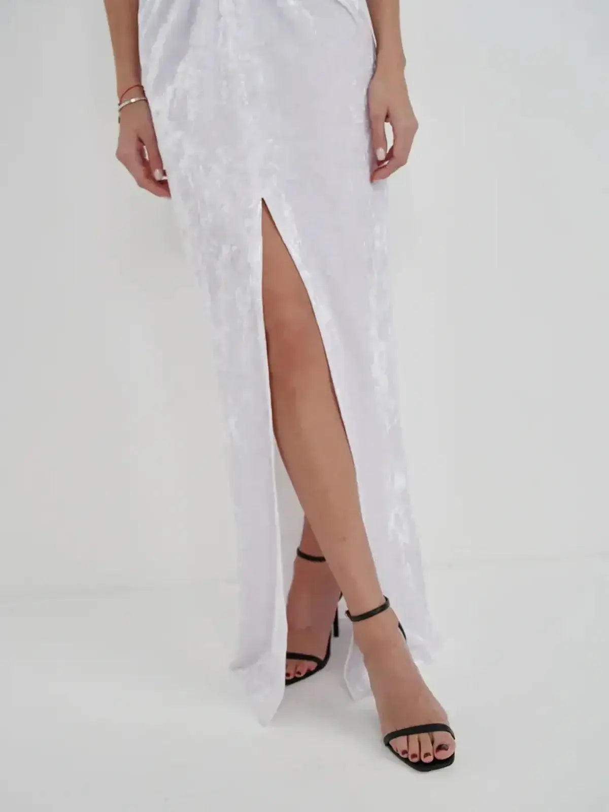 Close-up of a woman's legs in a sleek white crushed velvet maxi dress with a thigh-high slit and stylish black heels.