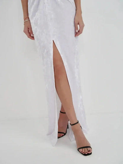 Close-up of a woman's legs in a sleek white crushed velvet maxi dress with a thigh-high slit and stylish black heels.
