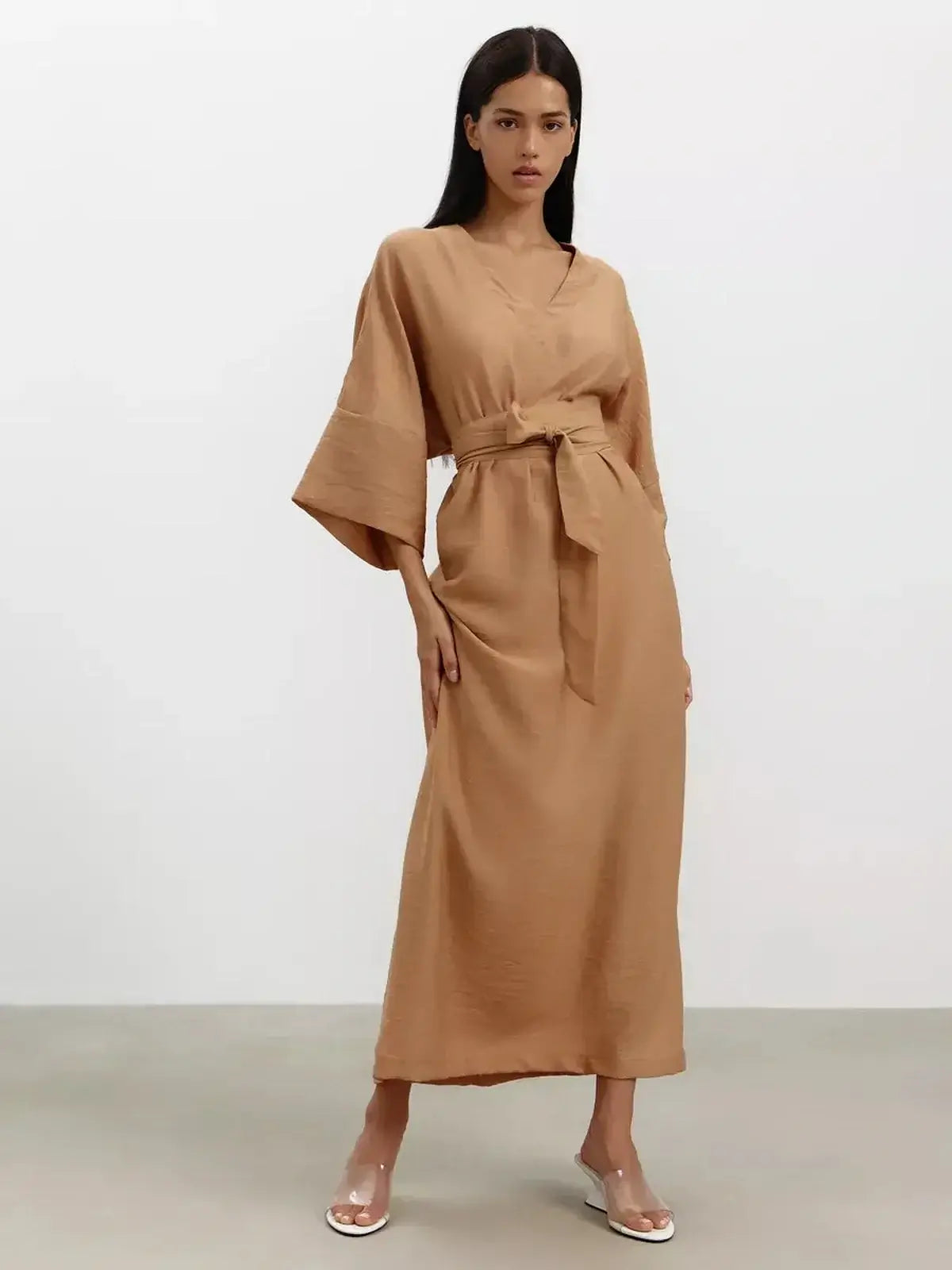 Model wearing a flowing maxi dress in a sandstone hue with wide sleeves and a detachable tie belt.