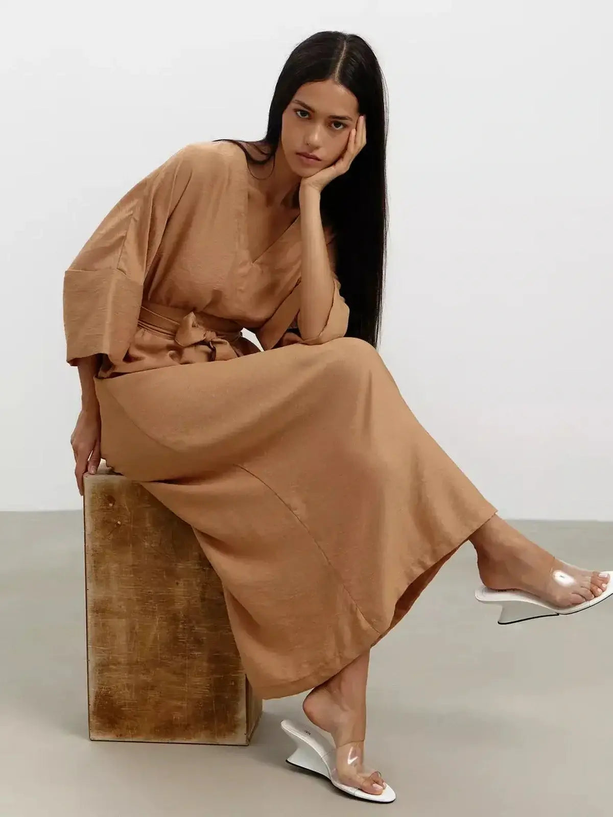 Model showcasing Maxi Dress Dune Whisper in sandstone hue with kimono sleeves and detachable belt, seated elegantly.