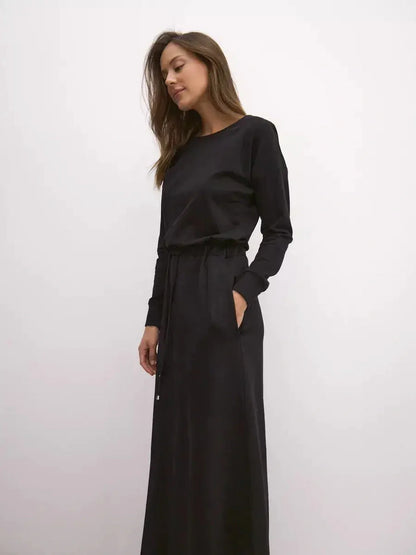 Model wearing a black maxi dress with long sleeves and cinched waist, showcasing urban elegance and comfort.