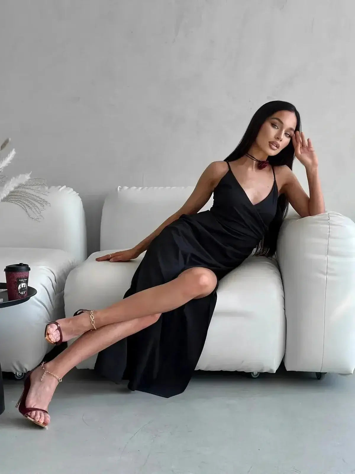 Woman in elegant black maxi dress Éclipse lounging on a sofa, showcasing a V-neckline and high slit.