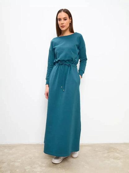 Model wearing a teal maxi dress with a cinched waist and long sleeves, showcasing elegant comfort and modern style.