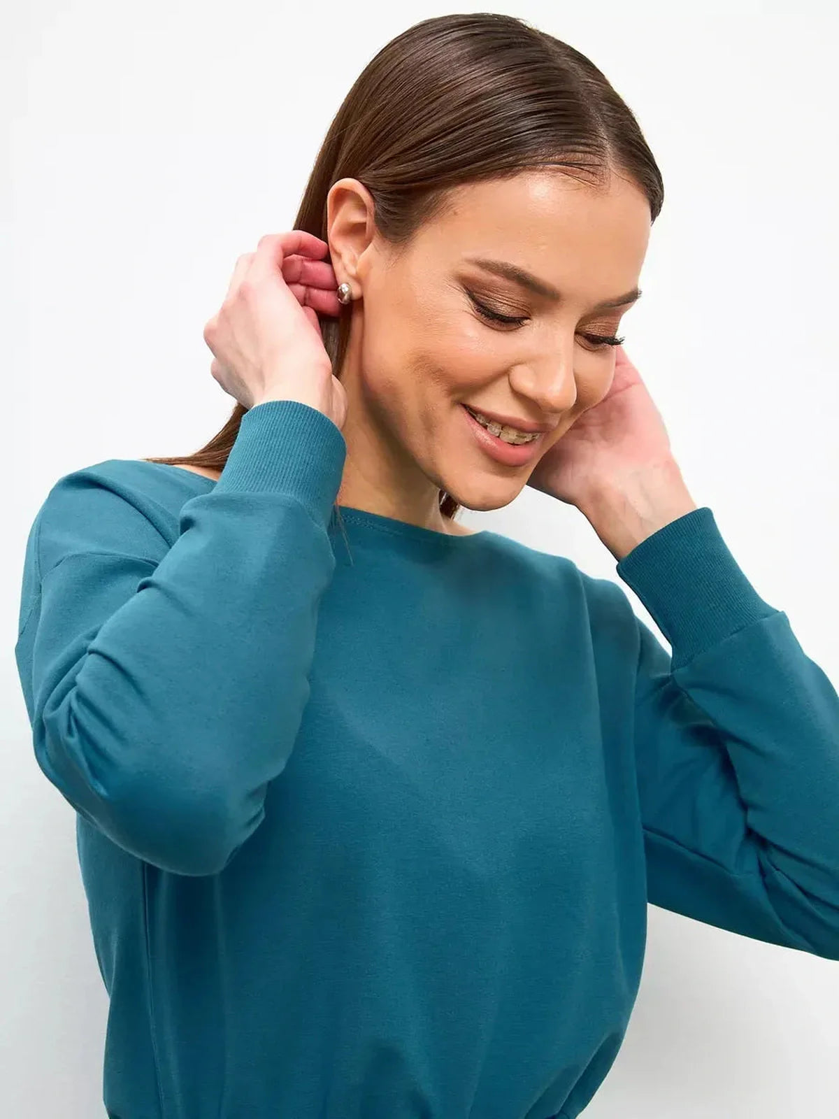 Smiling woman adjusting her earrings while wearing a teal long-sleeve top, showcasing elegance and comfort.