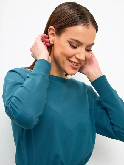 Smiling woman adjusting her earrings while wearing a teal long-sleeve top, showcasing elegance and comfort.